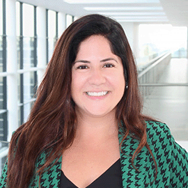 A smiling woman wearing a striped green blazer stands in front of a university corridor.